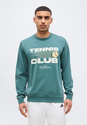 Giovane uomo che indossa una felpa turchese con la scritta "Tennis Club" e una grafica di una pallina da tennis, abbinata a pantaloni bianchi Adidas, su sfondo neutro.