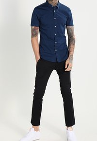Uomo con braccia tatuate che indossa una camicia a maniche corte blu navy con bottoni, pantaloni neri e sneakers bianche, in piedi con le mani nelle tasche.