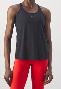 Czarny sportowy top na ramiączkach z zaokrąglonym dekoltem i cienkimi paskami, z subtelnym logo Nike. Noszony z jasnoczerwonymi legginsami.