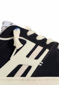 Sneaker in suede nero con dettagli in crema, caratterizzato da pelle testurizzata, lacci piatti e un design del logo prominente vicino ai lacci.