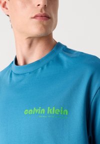 Τυρκουάζ βαμβακερό t-shirt με ριπ μπούστο, διακοσμημένο με πράσινη εκτυπωμένη λογότυπο "calvin klein" και επιπλέον κείμενο, λεία υφή και κοντά μανίκια.