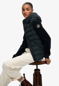 Doudoune noire sans manches avec capuche et devant texturé, associée à une manche noire ajustée. Un patch logo est présent sur le bras gauche. Pantalon de couleur naturelle porté.