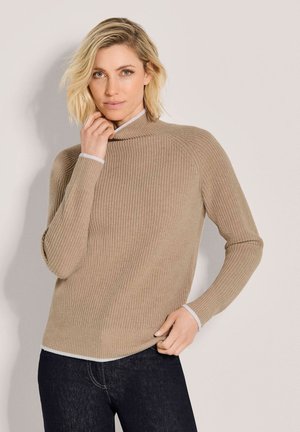 Vrouw met kort blond haar, gekleed in een beige geribde coltrui en donkere spijkerbroek, poseert met één hand bij haar gezicht en de andere op haar heup.