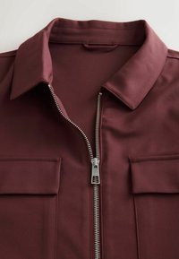 Burgundy zip-up shirt with a pointed collar, featuring two chest pockets and a silver zipper detailing along the front. Smooth fabric texture.