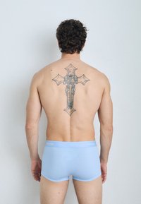 Boxer da uomo in cotone azzurro chiaro con una cintura morbida, indossati da un uomo con capelli ricci e un grande tatuaggio nero a forma di croce sulla schiena.
