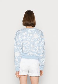 Svetle modrý květinový svetr s bílými vzory listů, kulatým výstřihem a žebrovanými manžetami. V kombinaci s bílými šortkami, pohled zezadu.