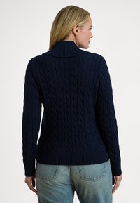 Sieviete ar blondiem matiem, nēsā zelta riņķa auskarus, tumši zilu pīto džempera džemperi un gaiši zilas džinsas, redzama no aizmugures uz balta fona.