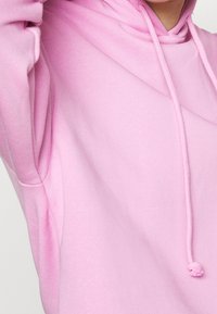 Sudadera con capucha rosa hecha de tela suave, con capucha ajustable mediante cordón y un corte holgado. La superficie tiene una textura lisa con detalles sutiles en las costuras.