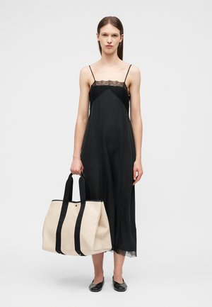 Γυναίκα με μαύρο φόρεμα-φόρμα που κρατά μια μεγάλη μπεζ τσάντα ώμου με μαύρες λαβές, στέκεται μπροστά σε ένα απλό φωτεινό φόντο.