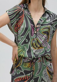Camicia a maniche corte di colore verde con audaci motivi a foglia multicolore, chiusura con bottoni frontale e vestibilità ampia, realizzata in tessuto leggero.