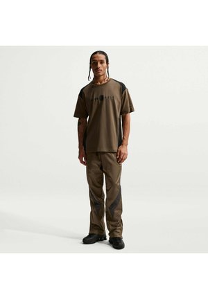 Mężczyzna z zaplecionymi włosami, ubrany w brązowo-czarną koszulkę Nike Air Shox oraz dopasowane spodnie i czarne trampki, stojący na jednolitym tle.