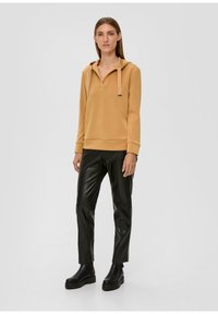 Pull beige clair avec capuche et cordons, associé à un pantalon en cuir synthétique noir ajusté et des bottes noires épaisses. Design simple.
