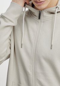 Lys beige hettegenser med glidelås laget av mykt stoff, med høy krage og snorer med metalliske topper, samt en frontlomme.