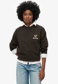 Felpa marrone con scollo rotondo a costine e vestibilità oversize, caratterizzata da ricami color crema "UNION MFG" e "Superdry". Collo bianco sottostante.