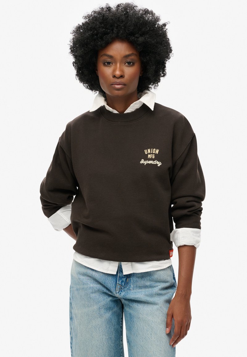 Felpa marrone con scollo rotondo a costine e vestibilità oversize, caratterizzata da ricami color crema "UNION MFG" e "Superdry". Collo bianco sottostante.
