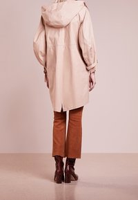 Femme debout, vue de dos, portant un manteau à capuche beige, un pantalon évasé marron et des bottes à talons marron foncé, sur un sol neutre.