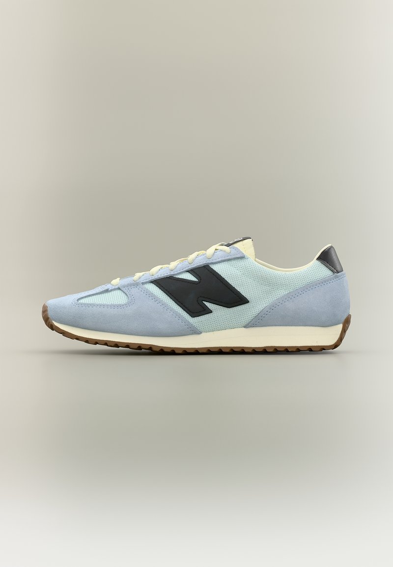 Baskets bleu clair et blanc avec logo noir sur le côté, lacets beige et semelle en caoutchouc marron, présentées en profil gauche sur fond neutre.
