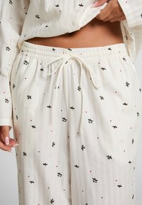 Wit gestreepte pyjamabeurt met knopen en een feestelijk hulstpatroon; bijpassende broek met elastische taille heeft een trekkoord en verspreidde rode accenten.