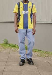 Gelbes und blaues vertikal gestreiftes Sporttrikot mit "Otokoc"-Logo, kombiniert mit hellblauen lockeren Hose und schwarzen Plateauschuhen.