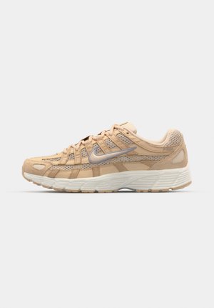 Zapatilla deportiva de color marrón claro con parte superior de malla, acentos de ante y una suela blanca texturizada. Presenta un logotipo plateado y cierre con cordones.