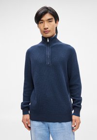Mørkeblå, ribbet strikket pullover med halvmund og høj krave. Har lange ærmer med strikkede manchetter og nederste kant.