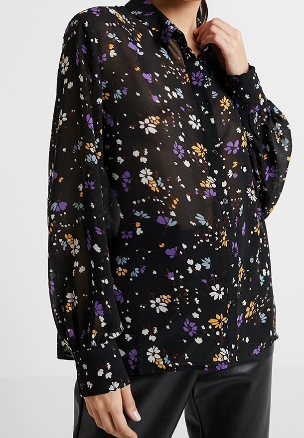 Blusa nera floreale con stampe colorate in viola, bianco e arancione. Tessuto trasparente con maniche lunghe e voluminose e chiusura a bottoni frontale.