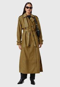 Olijfgroene leren trenchcoat met lange mouwen, dubbele knoopsluiting aan de voorkant, tailleriem en zijzakken. Gekozen in combinatie met zwarte jeans en laarzen.