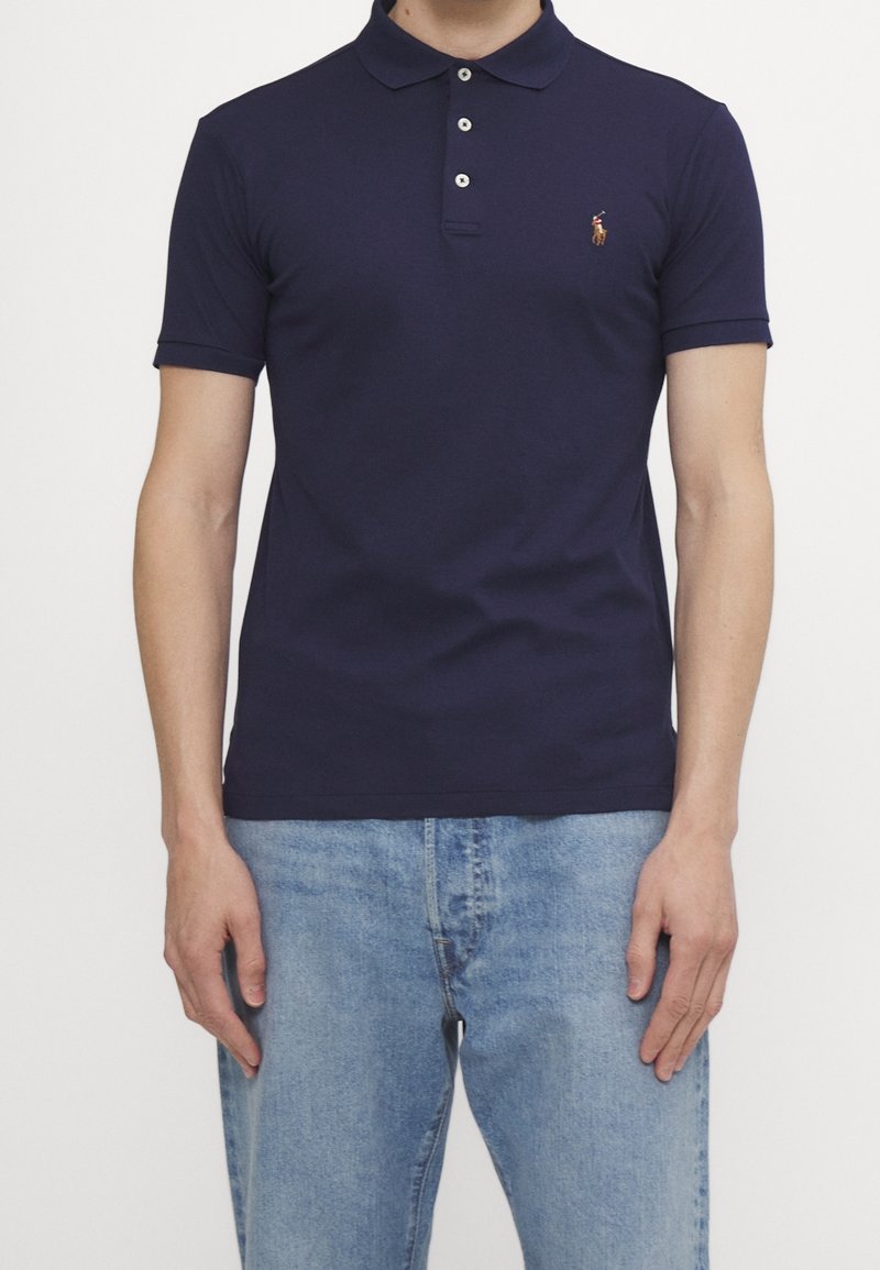 Homme portant un polo bleu marine ajusté avec trois boutons et un jean bleu clair sur un fond uni.