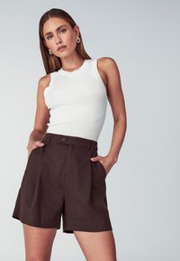 Witte geribbelde tanktop gecombineerd met hoogsluitende bruine shorts met zijzakken en een knoopsluiting, weergegeven tegen een neutrale achtergrond.