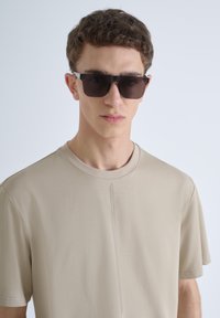 T-shirt beige con collo a giro e dettaglio di cucitura verticale. Modello che indossa occhiali da sole rettangolari scuri, con capelli corti e ricci.