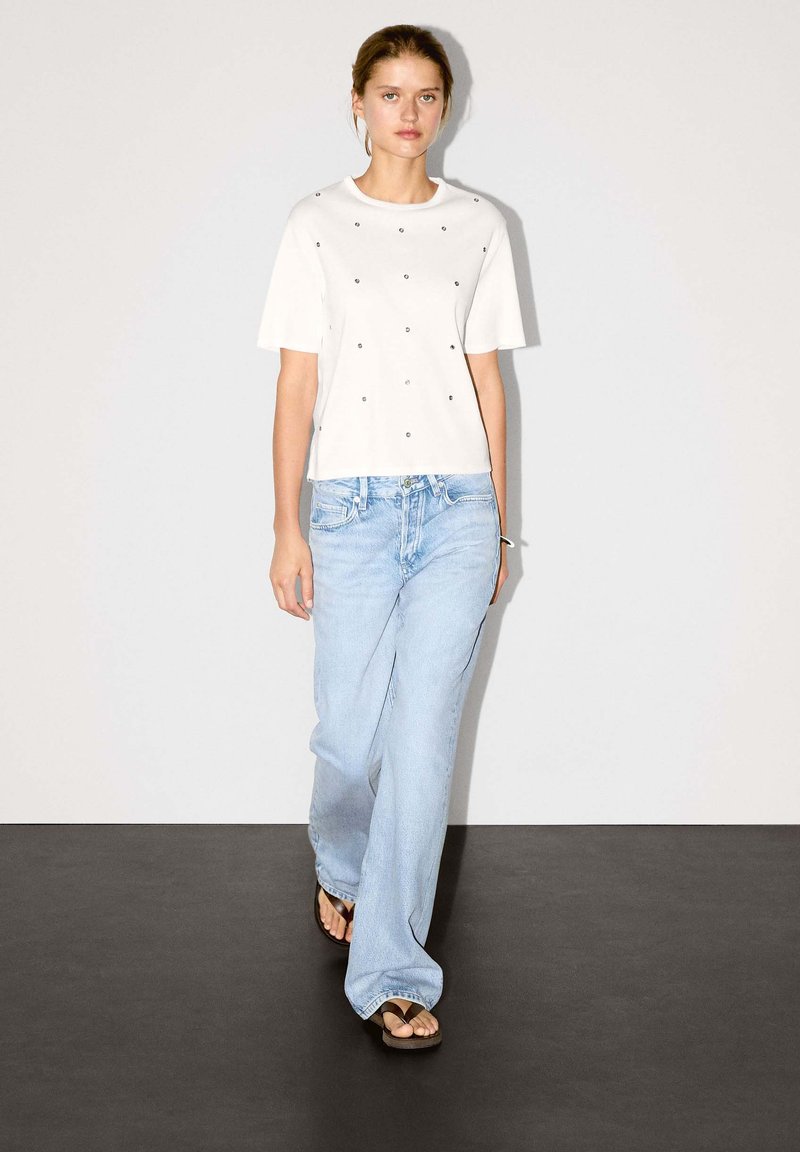 Wit T-shirt met korte mouwen en zwarte, edelsteenachtige versieringen, gecombineerd met lichtblauwe, loszittende jeans en bruine slippers.