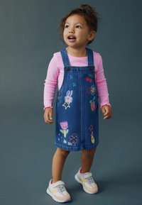 Vestito salopette in denim con disegni ricamati colorati, inclusi un coniglietto e dei fiori. Indossato sopra una maglia rosa a maniche lunghe, abbinato a scarpe da ginnastica.