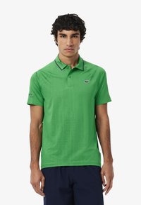 Polo de manga corta verde en tejido texturizado, con un sutil patrón geométrico, un cuello en contraste y un logotipo en la manga.