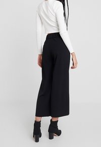 Pantalon noir à jambes larges avec une ceinture élastique, associé à un haut blanc à manches longues et court. Le mannequin porte des bottines noires.