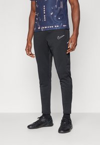 Schwarze Sporthose mit körperbetontem Design, seitlichen Streifen und kleinem weißen Nike-Logo. Getragen mit einem gemusterten Marineblauem Shirt und schwarzen Sneakers.