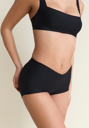 Kobieta w bezszwowej, czarnej bieliźnie z wysokim stanem oraz dopasowanym czarnym biustonoszu na jasnym, jednolitym tle.