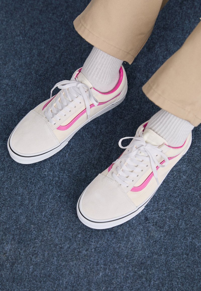 Zapatillas de ante blancas con acentos laterales rosas y suela negra. Cuentan con cordones blancos y calcetines blancos acanalados. Pantalones beige visibles sobre una superficie azul.