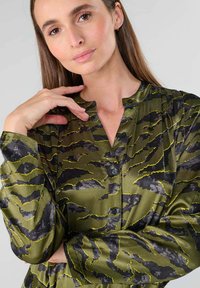 Grüne Satinbluse mit Wellenmuster in Schwarz und Gelb. Sie hat einen Knopfverschluss vorne und einen Stehkragen sowie lange Ärmel.