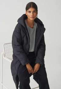 Schwarze Steppjacke mit Kapuze, im Rautenmuster, mit Reißverschluss vorne, getragen über einem grauen T-Shirt, kombiniert mit dunklen Hosen.