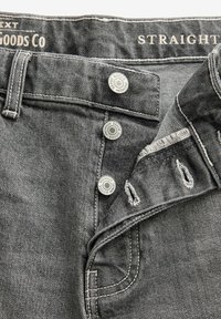 Graue Denim-Jeans im Straight-Cut mit silbernen Knopfverschlüssen, Kontrastnähten und fransigen Details am Bund. Das Etikett trägt die Aufschrift "Premium Denim Goods Co."