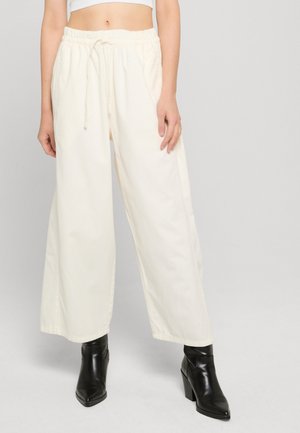 Osoba ubrana w luźne, szerokie spodnie w kolorze kremowym z wiązaniem w talii oraz czarne, szpiczaste botki, stojąca na szarym tle.