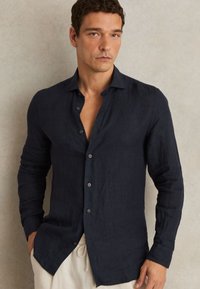 Hombre con camisa azul marino oscura abotonada de manga larga y pantalones beige, de pie frente a una pared de color neutro.