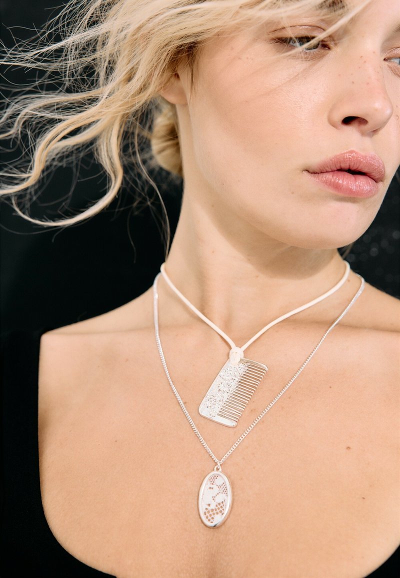 Femme blonde aux cheveux lâchés porte deux colliers en argent, l'un avec un pendentif en forme de peigne et l'autre avec un pendentif ovale à motif.