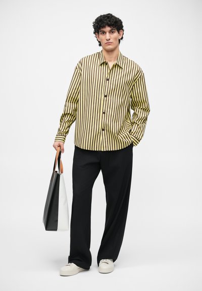 Gestreifte Button-Up-Bluse in Gelb und Braun, kombiniert mit lockeren schwarzen Hosen und weißen Sneakers. Das Model hält eine schwarz-weiße Tragetasche.
