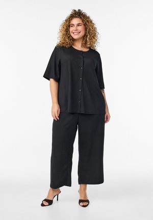 Hymyilevä nainen, jolla on kiharat hiukset ja päällään löysä musta napitettava yläosa, leveälahkeiset housut ja avokärkiset korkokengät, seisoo valkoisen taustan edessä.