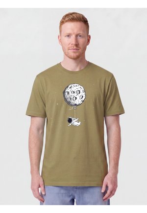 Homme portant un t-shirt vert olive avec un graphisme d'astronaute tenant un ballon en forme de lune flottant dans l'espace, debout devant un fond uni.
