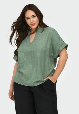 Frau mit langen dunklen Haaren, die eine lockere, hellgrüne Kurzarmbluse und schwarze Hose trägt, posiert mit einer Hand in der Tasche.