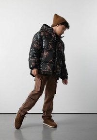 Chłopiec w brązowej czapce, czarnej kurtce puchowej w wzory, brązowych spodniach cargo i brązowych sneakersach, pochylony na jednej nodze wewnątrz budynku.