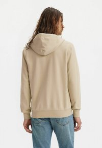 Beige Kapuzenpullover mit Kordelzug, weichem Material und langen Ärmeln. Verfügt über eine Kängurutasche und gerippte Bündchen. Getragen mit blauen Jeans.