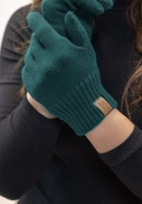 Türkise gestrickte Handschuhe mit gerippten Bündchen und einem kleinen, braunen Etikett, das angenäht ist, präsentiert auf einer Hand vor einem schwarzen Langarmoberteil.